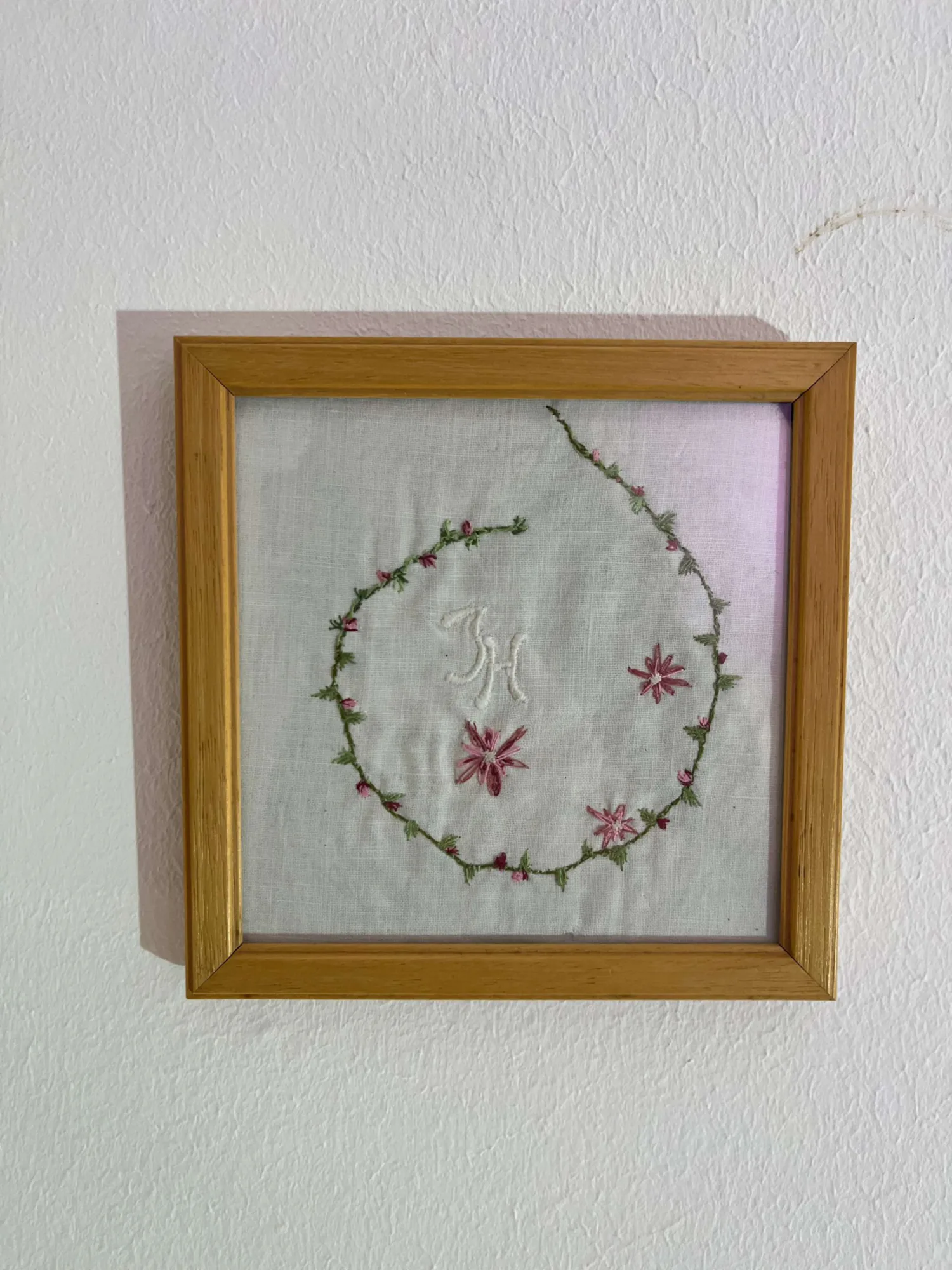 Das kunstwekr zeigt ein in Holz gerahmten Stoff auf dem eine feine Blumenranke in Kreisform gestickt ist. In der Mitte wurden die Initialen JH gestickt.