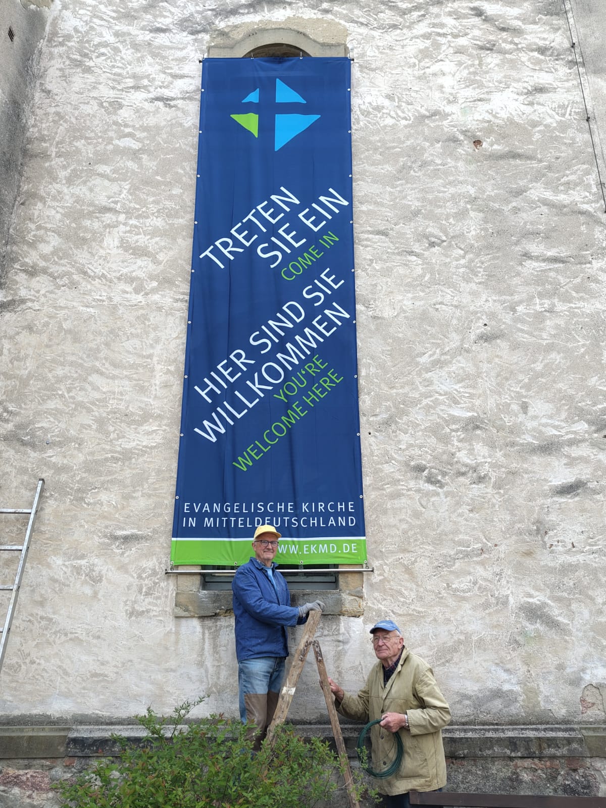 Banner der Kirche mit der Aufschrift: Treten Sie ein. Hier sind die willkommen. Davor stehen 2 Männer, die es vermutlich angebracht haben.