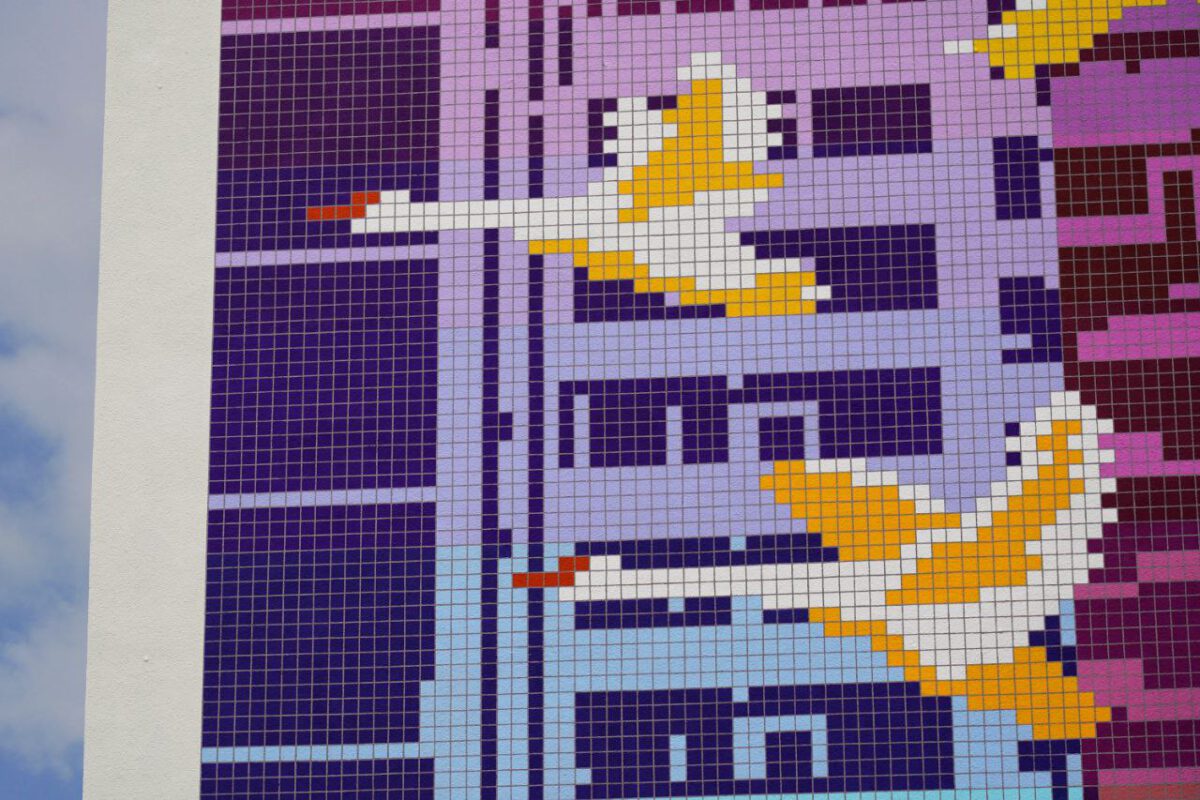 Mural 'Flight of the Swans', colored rectangular tiles create a pixelish Image of white-yellow flying swans, in front of a modern building facade. in blue-violet colors.
